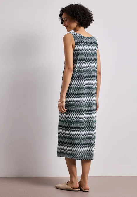 Femme en robe longue à motif zigzag, sandales aux pieds, posant devant un mur blanc.