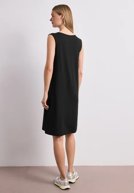 Frau von hinten in ärmellosem schwarzem knielangem Kleid und weißen Sneakern vor hellem Hintergrund.