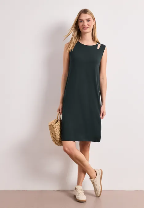 Femme en robe noire épurée sans manches à découpe épaule, sac en paille et sandales blanches.