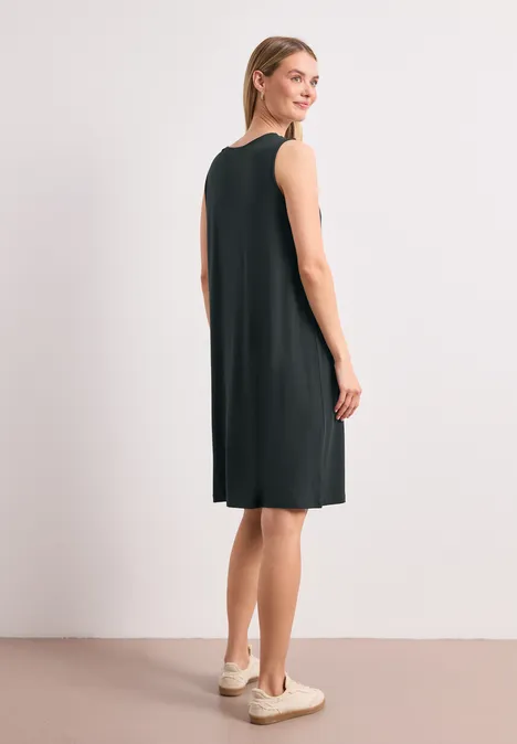 Une femme porte une robe noire sans manches jusqu'aux genoux et des baskets, vue de côté.