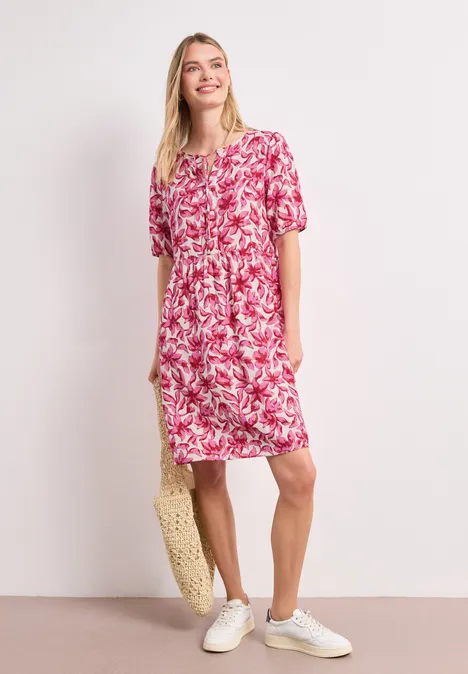 Frau trägt locker sitzendes rosa-weißes Blumenkleid, weiße Sneaker, Strohtasche