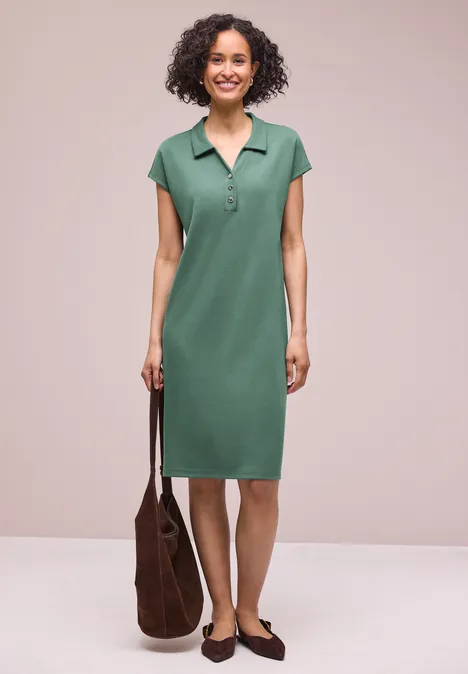 Femme en robe polo verte devant fond neutre, tenant un sac marron et portant des mocassins.