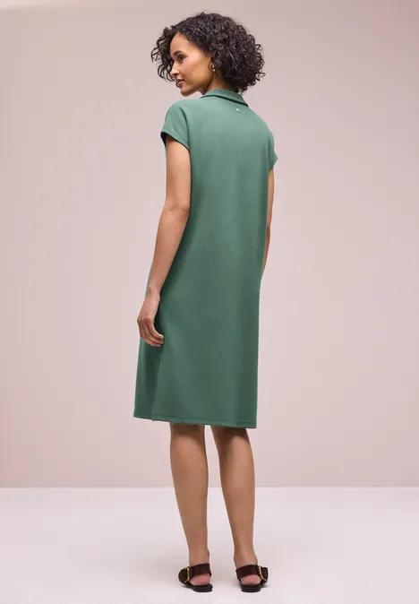 Une femme vue de dos porte une robe polo verte jusqu’aux genoux, manches courtes, et sandales noires.