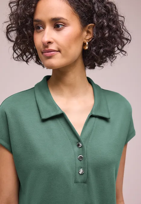 Femme aux cheveux bouclés porte un polo vert à manches courtes avec col et patte de boutonnage.