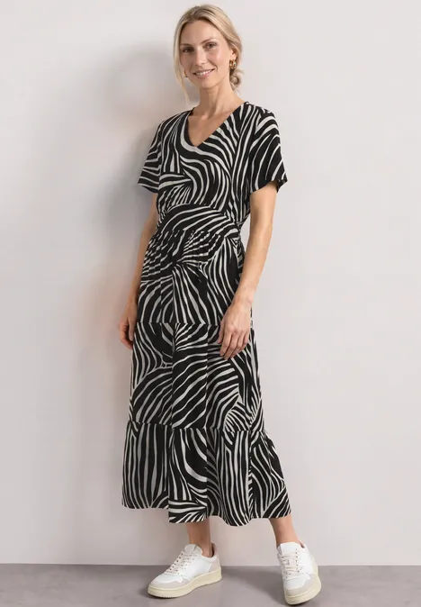 Frau in schwarz-weißer Zebra-Midikleid und weißen Sneakern vor hellem Hintergrund.