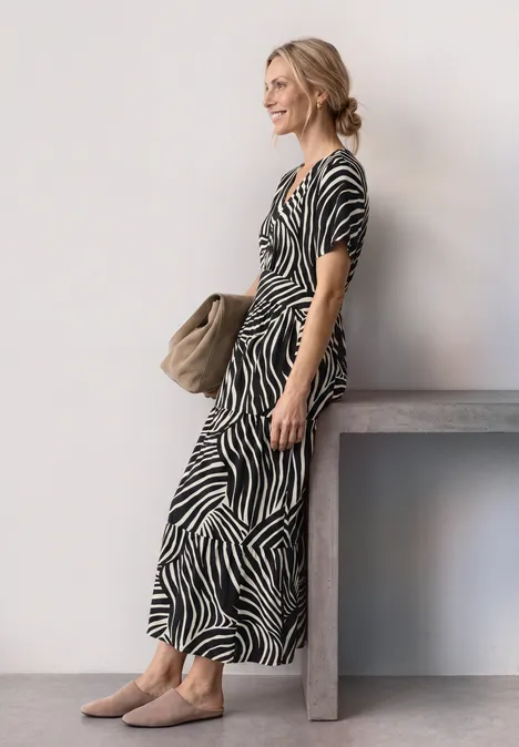 Frau im Zebra-Midikleid mit Tasche, lässig an einem Tisch vor heller Wand gelehnt.