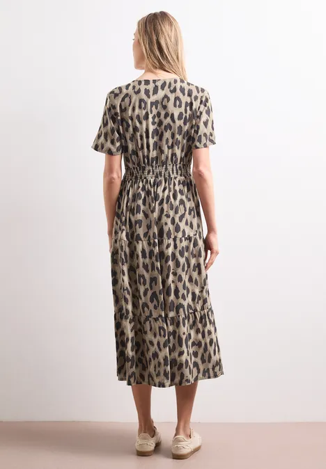 Rückansicht einer Frau in Midi-Kleid mit Leopardenmuster, kurzen Ärmeln und beigen Schuhen vor weißer Wand.