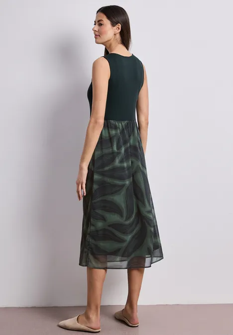 Frau von hinten in ärmellosem dunkelgrünem Oberteil und gemusterter, grün-schwarzer Midi-Rock, heller Hintergrund.