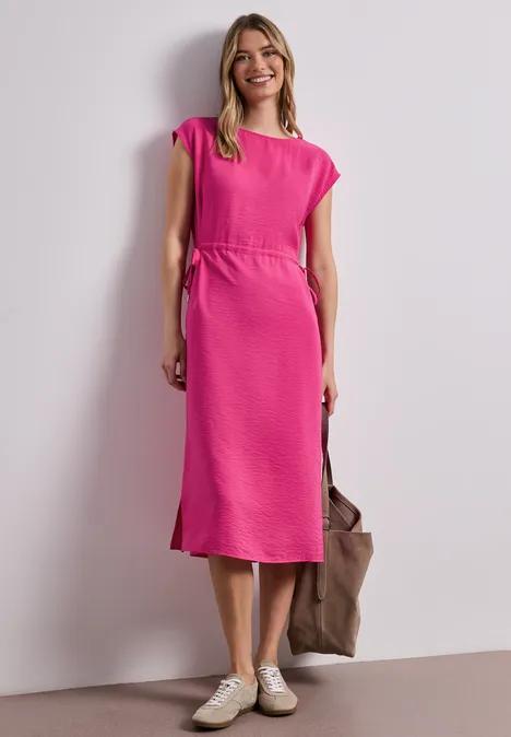 Eine Frau trägt ein pinkes Midi-Kleid, weiße Sneaker und hält eine braune Tasche.