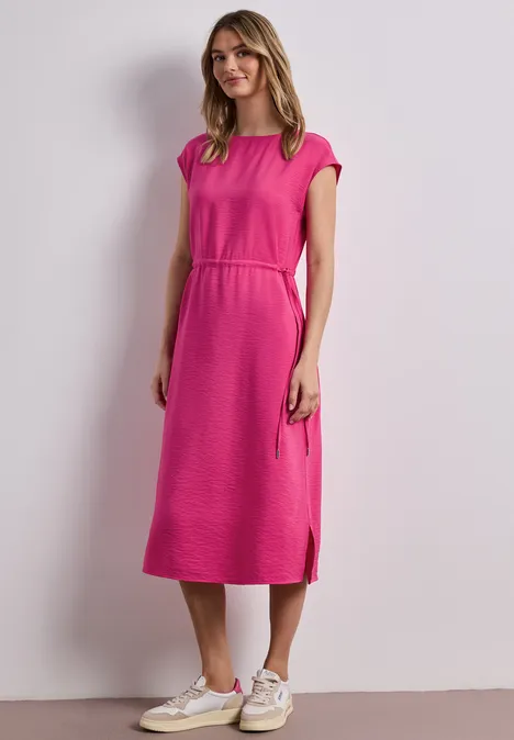 Frau in knielangem pinken Kleid und weißen Sneakers vor hellem Hintergrund.