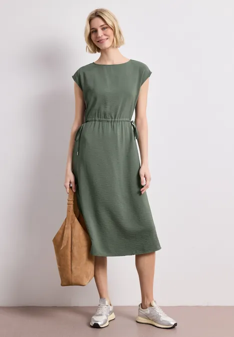 Femme en robe midi verte cintrée, baskets blanches et sac cabas brun.