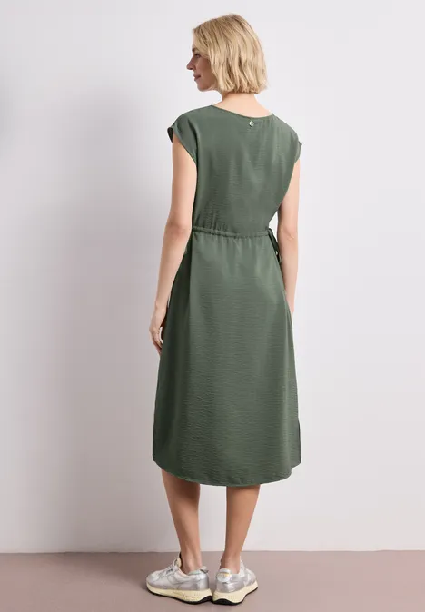 Une femme de dos porte une robe midi kaki sans manches et des baskets blanches, sur fond neutre.