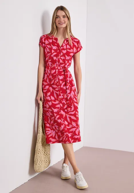 Vrouw draagt rode midijurk met wit bladpatroon, V-hals, tailleceintuur, witte sneakers en netzak.