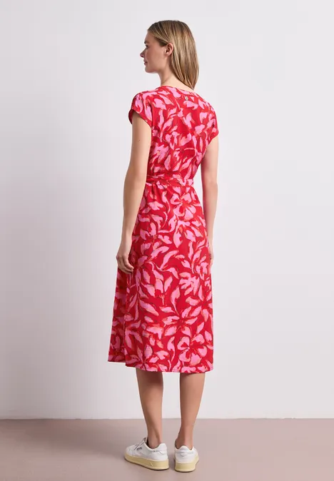Vrouw in profiel draagt een rode midijurk met wit bladmotief en witte sneakers voor een eenvoudige achtergrond.