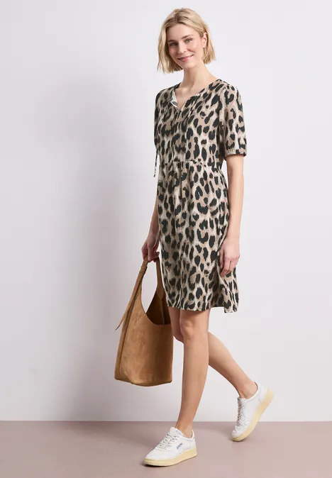 Frau in knieumspielendem, kurzärmeligem Leopardenkleid mit weiß-gelben Sneakern und braunem Veloursbeutel.