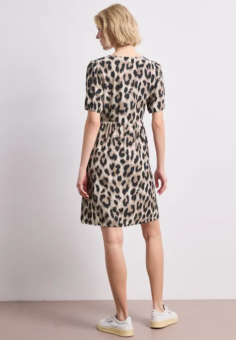 Frau von hinten in kurzem, leo-getupftem Kleid mit kurzen Ärmeln und hellen Sneakern vor neutralem Hintergrund.