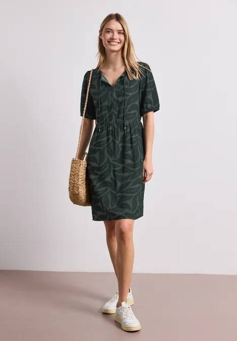 Frau in dunkelgrünem, kurzärmligem Hemdblusenkleid mit Blattmuster, weißen Sneakern und geflochtener Strohtasche.