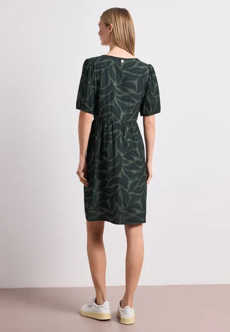 Frau von hinten in grünem Kleid mit Blattmuster und weißen Sneakern vor weißem Hintergrund.