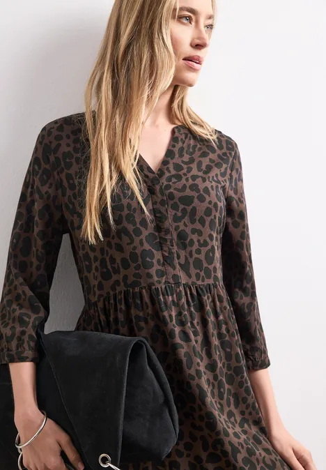 Frau in braunem Leo-Kleid, 3/4-Ärmel, hält schwarzen Wildleder-Clutch, langes blondes Haar.