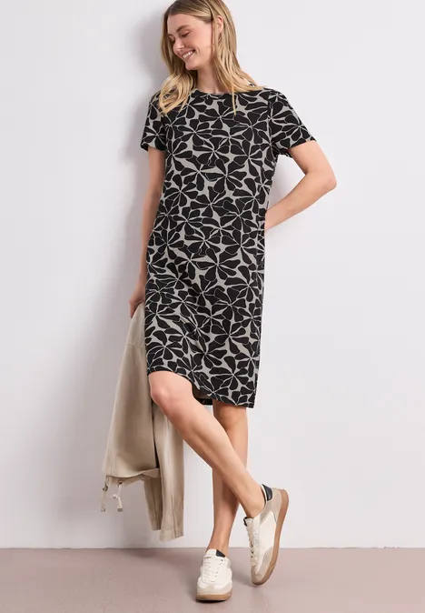 Frau in lockerem, knielangem schwarz-weißem Kleid mit kurzen Ärmeln, weißen Sneakern und heller Jacke.