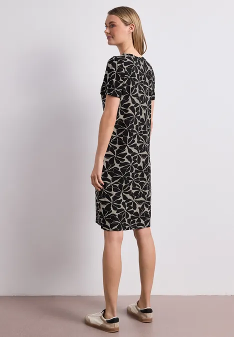 Frau steht seitlich vor weißer Wand, trägt schwarzes Kleid mit weißem Blattmuster und Slip-on-Sneakers.