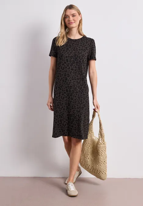 Frau in schwarzem, gemustertem Kurzarmkleid mit Strohtasche und weißen Slippern.