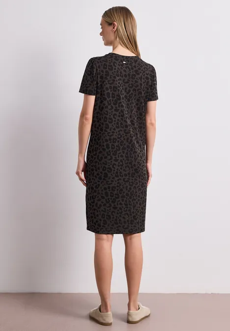 Rückansicht einer Frau in knielangem, schwarzem Kleid mit dezentem Muster und kurzen Ärmeln vor neutralem hellem Hintergrund.