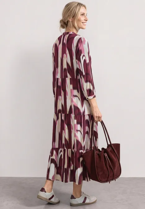 Midi-Kleid mit Split Neck im Ethno-Look rustic rouge Frau von hinten in lockerem Maxikleid mit abstraktem Muster, trägt eine burgunderfarbene Tasche und Sandalen.