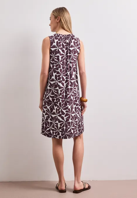 Frau von hinten in einem ärmellosen, lila Kleid mit weißem Blumenmuster, trägt Sandalen.