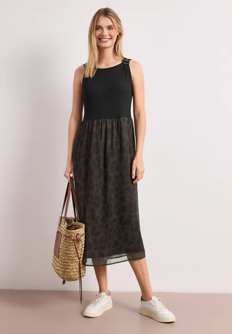 Een vrouw in een mouwloos zwart topje, een midi rok met print, witte sneakers en een gevlochten tas