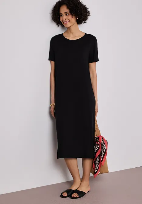 Frau in schlichtem schwarzem Midikleid mit kurzen Ärmeln und Sandalen, hält eine bunte Tasche.