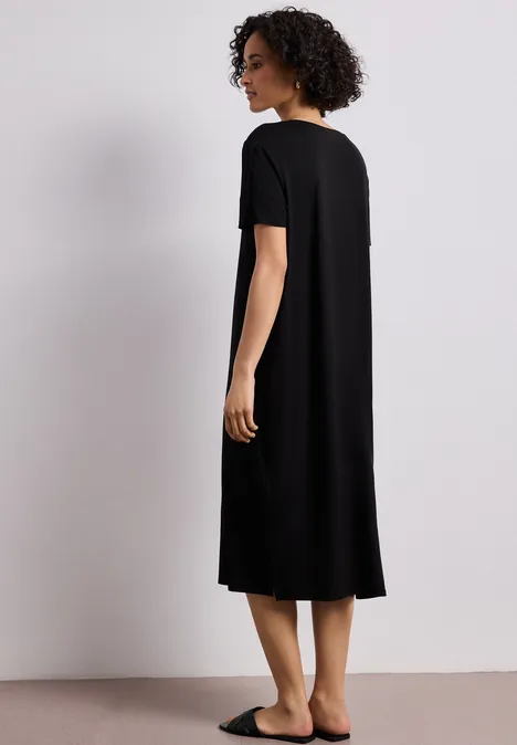 Eine Frau trägt ein schwarzes, knielanges T-Shirt-Kleid und steht seitlich vor neutralem Hintergrund.