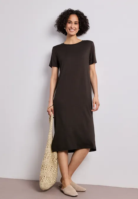 Person trägt schlichtes braunes T-Shirt-Kleid, geflochtenen Shopper und bequeme beigen Slipper.