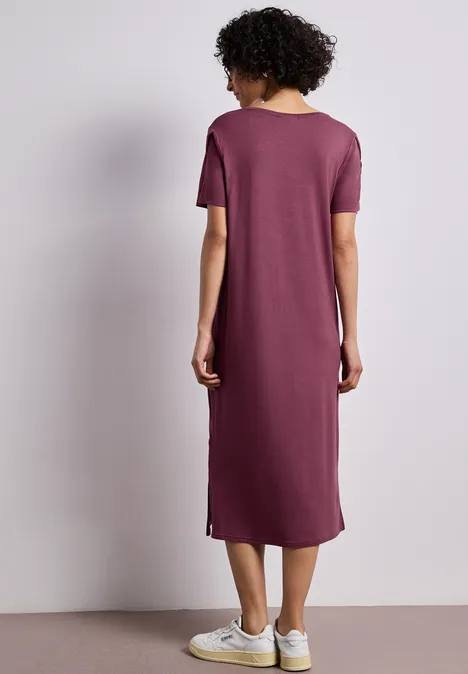 Rückenansicht einer Frau in schlichtem lila Midikleid und weißen Sneakers vor schlichtem Hintergrund.