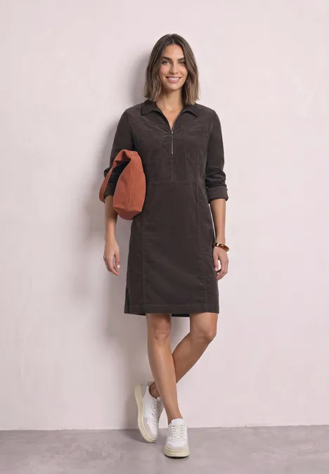 Frau in dunkelbraunem Cordkleid mit halbem Reißverschluss, weißen Sneakern und orangem Clutch.