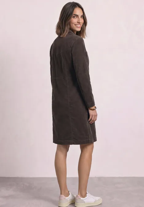 Frau steht seitlich mit Blick über die Schulter, trägt braunes knielanges Kleid und helle Sneaker.