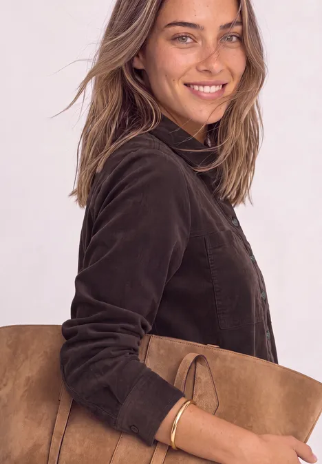 Lächelnde Frau mit brauner Wildledertasche und dunkler Jacke schaut in die Kamera.