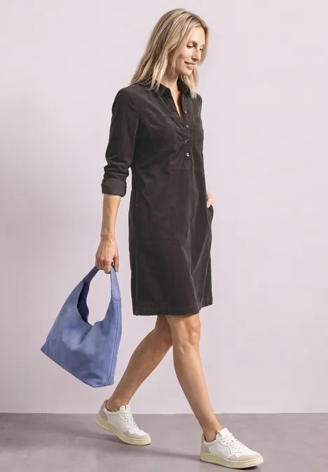Une femme blonde porte une robe en velours gris foncé, des baskets blanches et un sac bleu.
