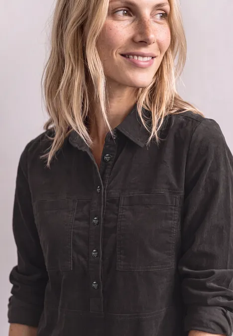 Une femme blonde sourit, porte une chemise en velours côtelé sombre, regard de côté.