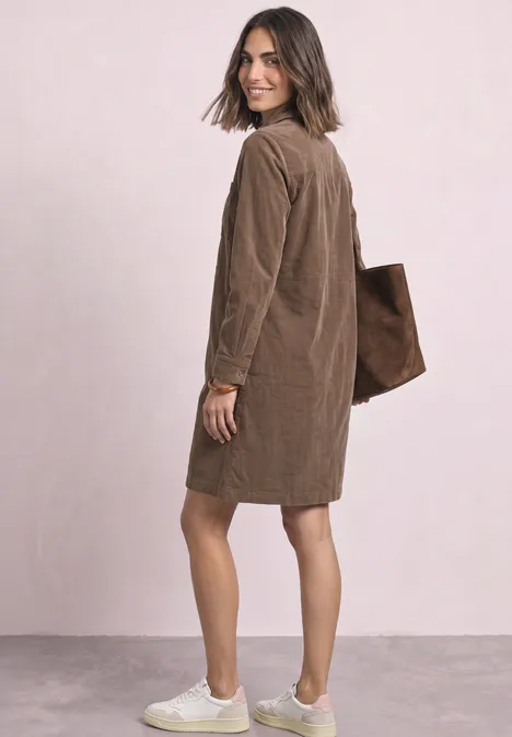 Knielanges Kleid aus softem Cord velvet mocha Frau in braunem Cordmantel und weißen Sneakern trägt eine Tote Bag, schaut lächelnd über die Schulter.