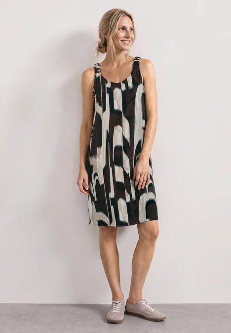 Robe mi-longue à bretelles froncées espresso brown Frau in lockerem, schwarz-weiß gemustertem ärmellosem Kleid und grauen Schnürschuhen vor hellem Hintergrund.