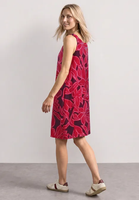 Robe mi-longue à bretelles froncées salsa red Femme de profil portant une robe sans manches rouge à motif floral rose et des chaussures blanches.
