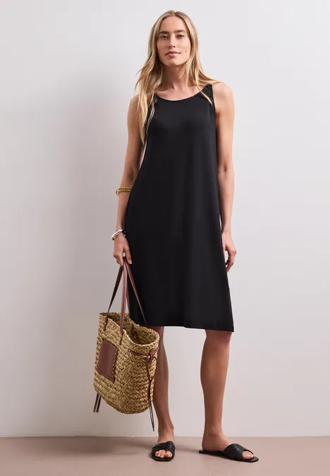 Eine Frau trägt ein schwarzes ärmelloses Kleid, hält eine geflochtene Tasche und trägt schwarze Sandalen.
