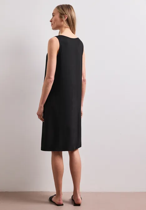 Frau von hinten in ärmellosem, knielangem schwarzen Kleid und flachen Sandalen vor neutralem Hintergrund.