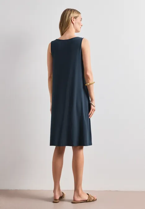 Rückenansicht einer Frau in ärmellosem dunkelblauen Kleid und flachen Sandalen vor heller Wand.