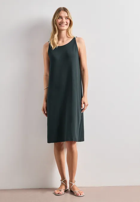 Knielanges Kleid mit Schnallendetail deep bound green Frau in schlichtem, ärmellosem, knielangem dunkelgrünem Kleid und Sandalen vor hellem Hintergrund.