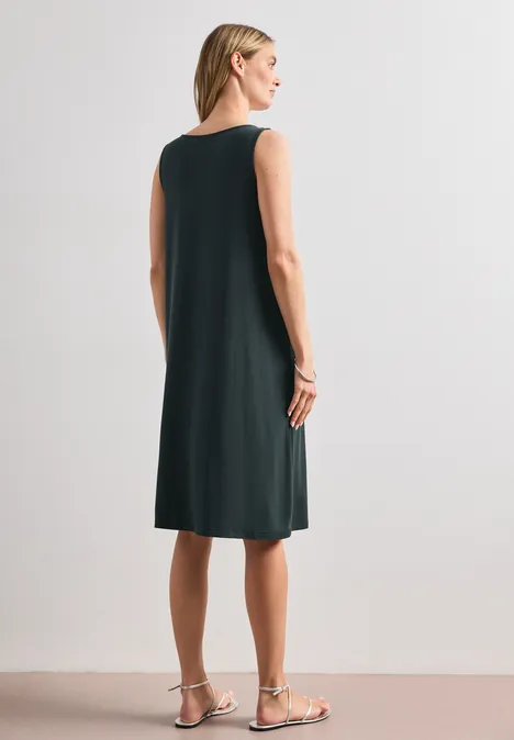 Knielanges Kleid mit Schnallendetail deep bound green Frau von hinten in ärmellosem, knielangem, dunklem Kleid und flachen Sandalen vor hellem Hintergrund