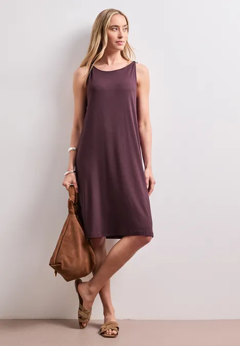 Frau in locker fallendem, ärmellosem violettem Kleid, mit brauner Tasche und Sandalen vor weißem Hintergrund.