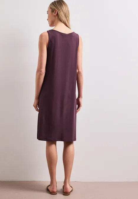 Rückenansicht einer Frau in ärmellosem, knieumspielendem, dunkellila Kleid und flachen Sandalen.
