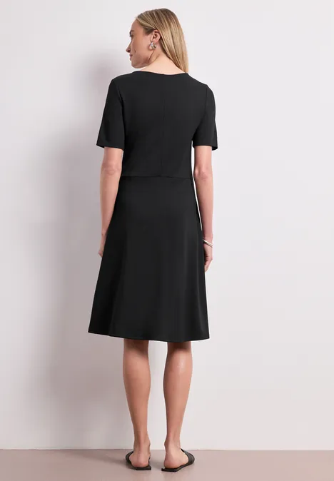 Robe en jersey à manches courtes Black Femme de dos portant une robe noire mi-longue, manches courtes et chaussures plates devant un fond clair.